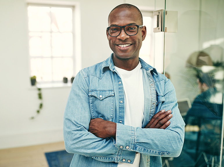 Smiling man with glasses standing confidently in a modern office with arms crossed.
