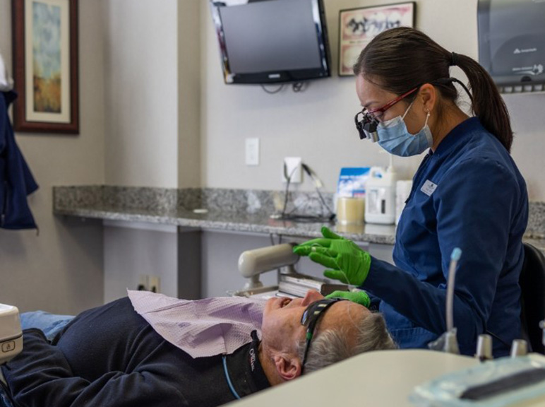 Operating Microscope Dental hygienist treating a patient in a dental exam room while wearing protective gear.